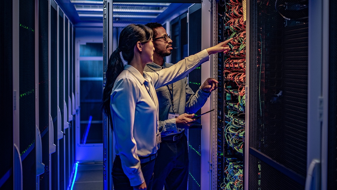 Two employees in a data center pointing at ethernet cables on the back of a rack of networking equipment. 