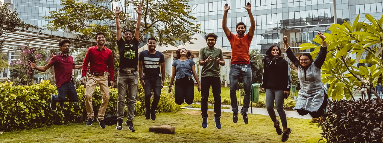 Nine people jumping mid-air outside with a building in the background.