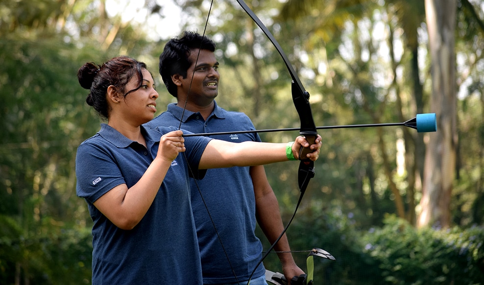 Two people stand together in nature wearing Cisco shirts while one uses a bow and arrow.