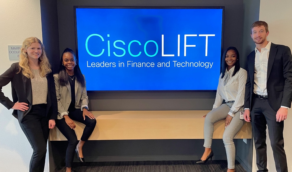 Four people huddle by a TV screen that reads “Cisco LIFT. Leaders in Finance and Technology.”