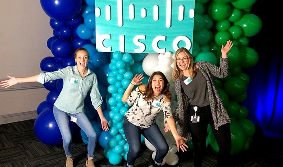 Three people pose with balloons and a Cisco sign in the foreground.