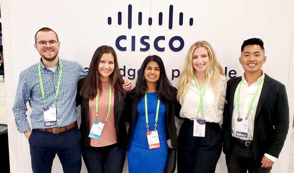 Group of five people stand together wearing lanyards with the Cisco logo in the background.