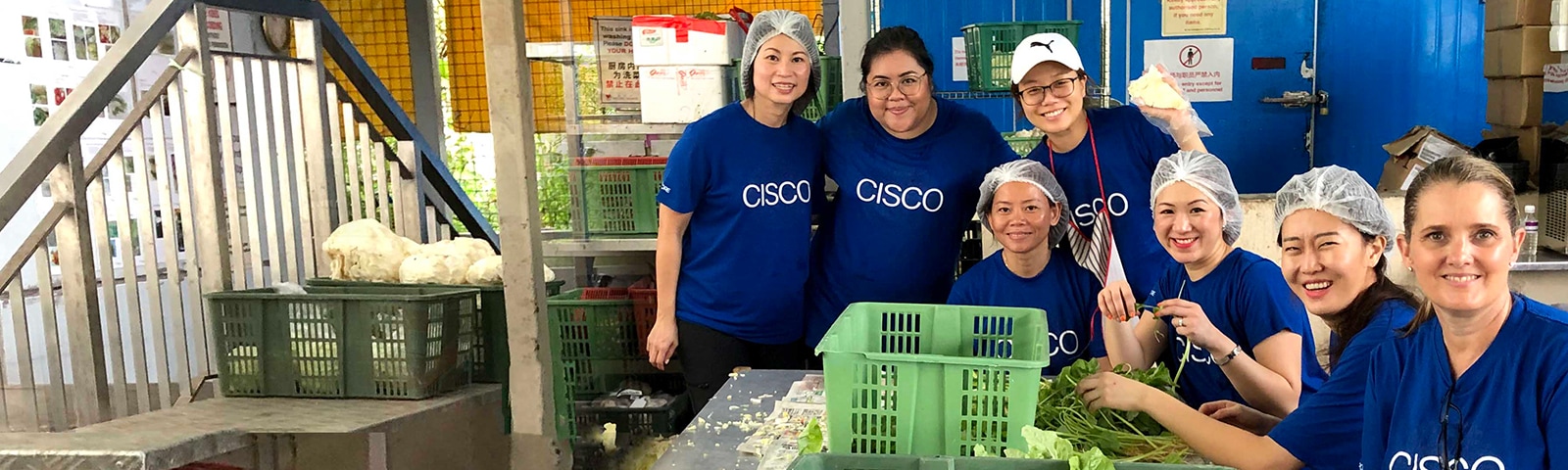 Seven women wearing the same blue Cisco shirt volunteering at a food bank while sorting lettuce