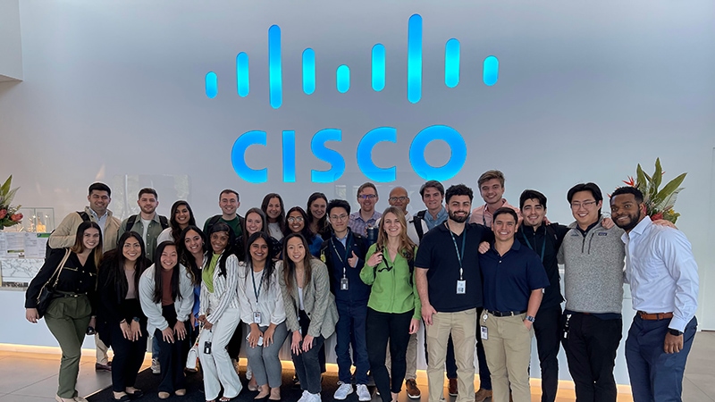 A large cohort of Ciscos Finance University Programs stands in front of a Cisco sign. 