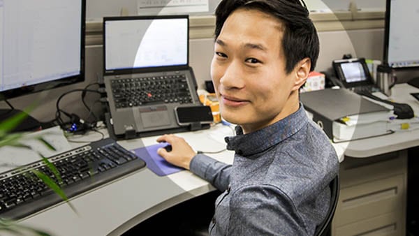 A smiling Asian man looking at you in a grey shirt working at his desk in front of a keyboard, a laptop, and a monitor.