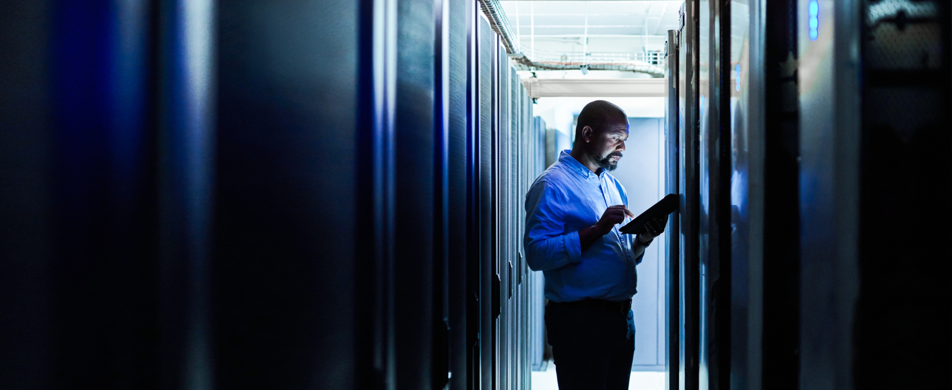 IT worker walking through the enterprise data centre checking network dashboards on a tablet.