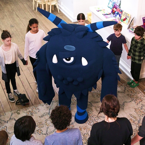 A group of children standing in a circle around a large blue monster mascot in a brightly lit classroom with books and toys on display.