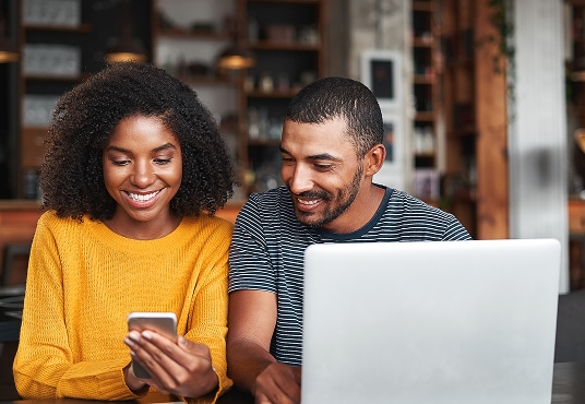 Couple sitting at a table in a cafe, looking at a smartphone together; one is holding the phone and the other is sitting in front of a laptop.