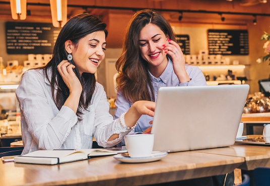 Two young businesswomen sitting in cafe at table, looking at a laptop together.