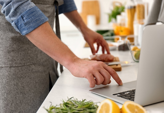 Person wearing an apron using a laptop in a kitchen, with fresh ingredients like lemons and herbs on the counter.