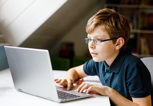Young kid with glasses learning at home on a laptop for school.
