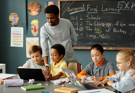 Diverse group of primary school children using laptops in pairs to study, sitting at a large desk in a classroom while their teacher assists them.