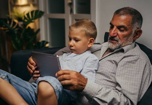 Grandparent and their grandchild relaxing on sofa at home using a tablet computer.