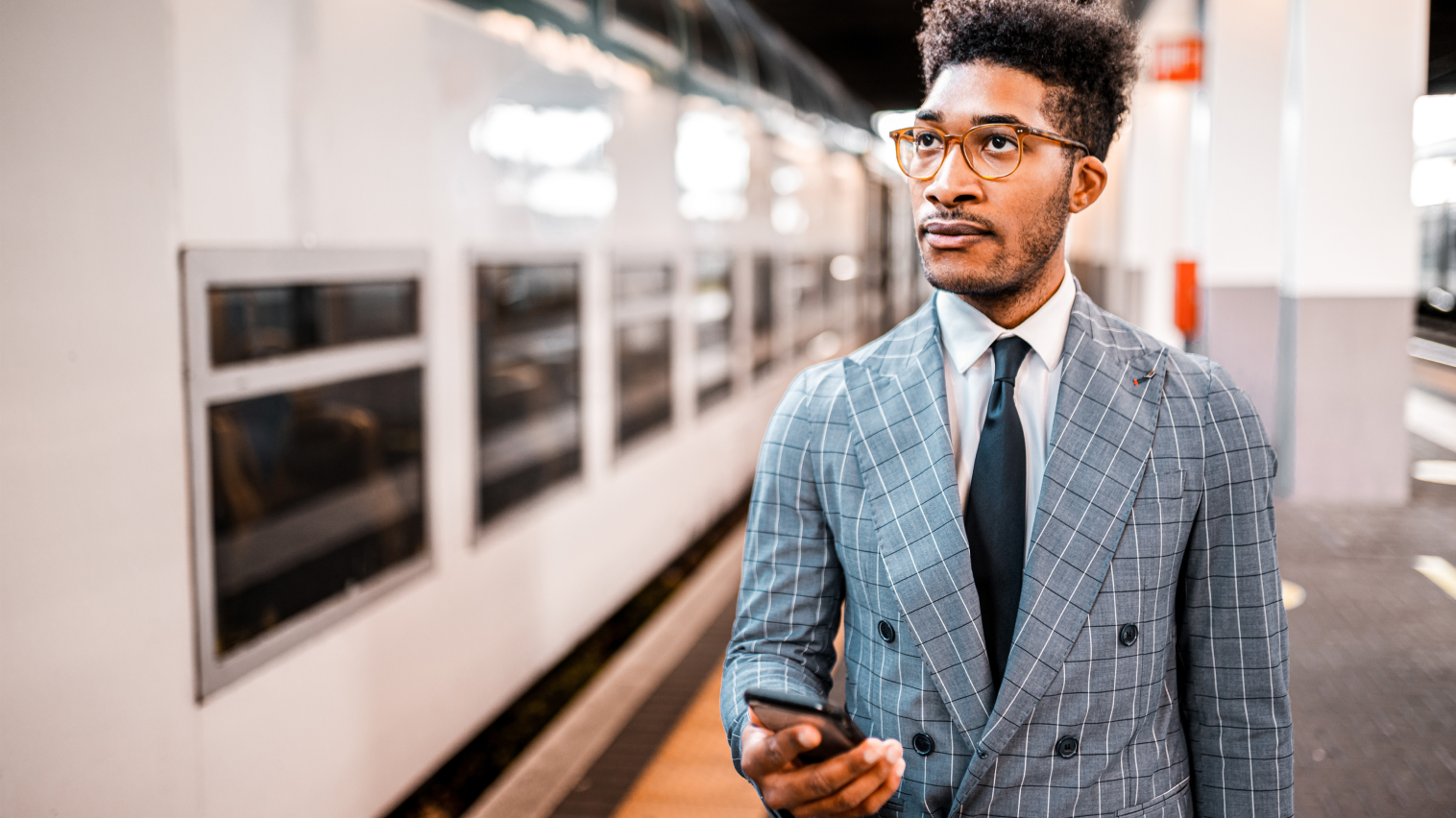 A man wearing red-rimmed glasses, suit and tie standing in a subway holding a smartphone.