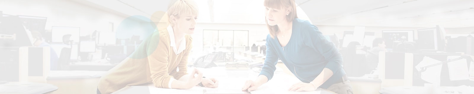 A woman in a yellow collared sweater brainstorming over a tablet with another woman in a blue sweater on a communal table in a work office setting.