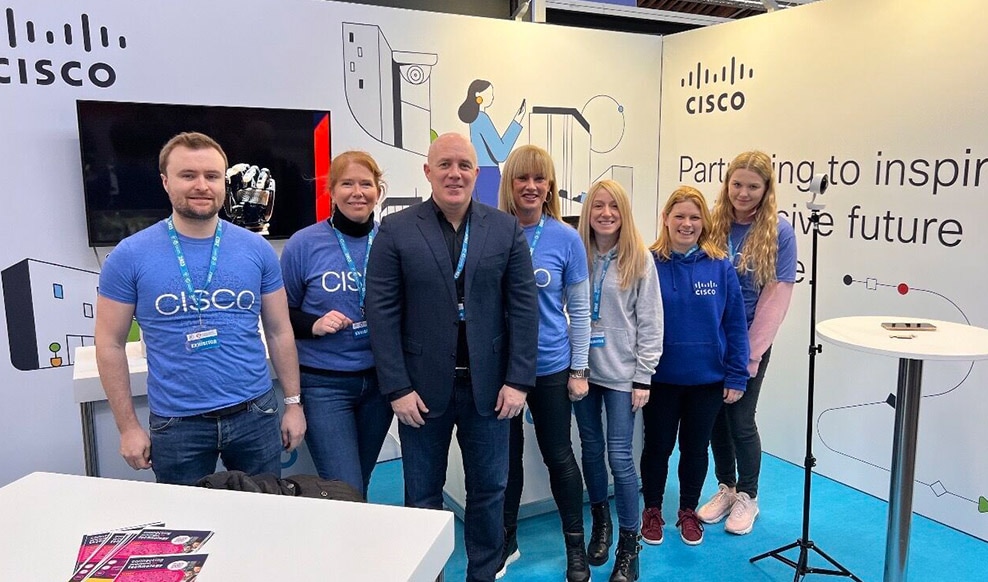 Three people smile and pose together with Cisco hardware in the background.