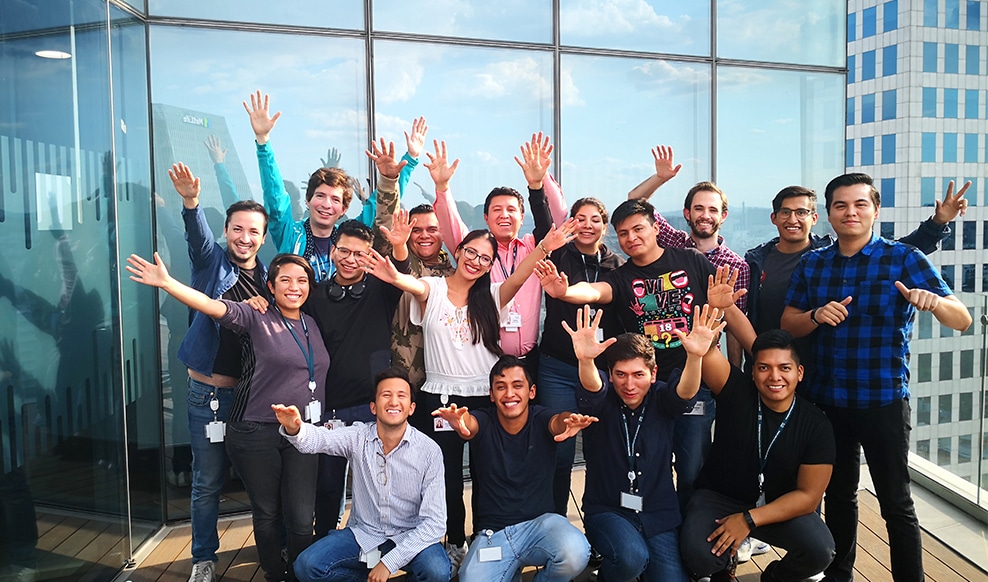 Group of sixteen people raise their arms and smile on a building terrace.