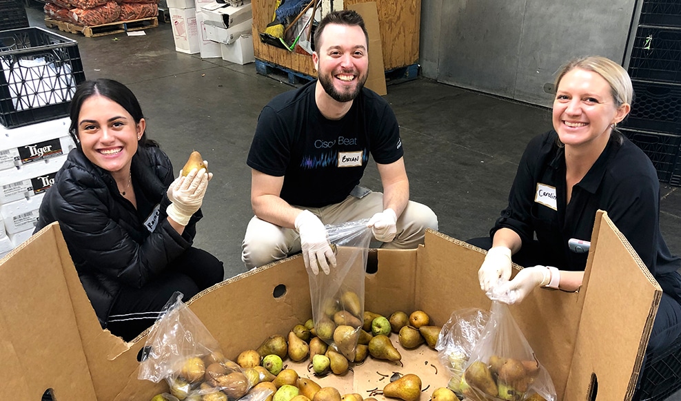 Three people smile while packing pears into plastic bags.