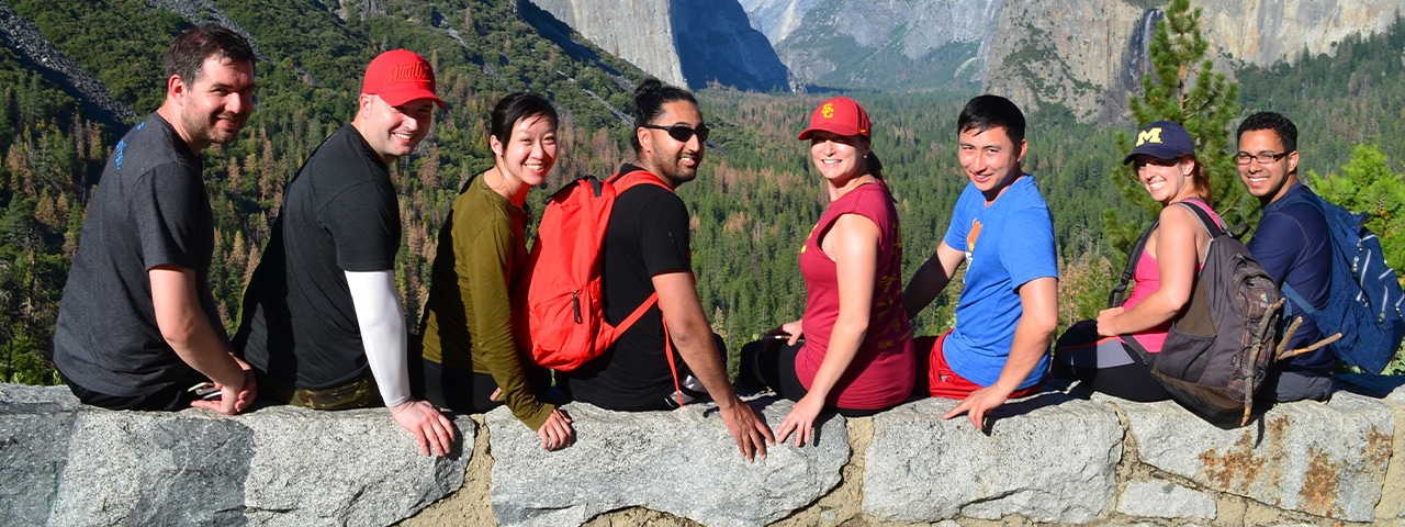Group of people sit on rock ledge with foliage and mountains in the background.