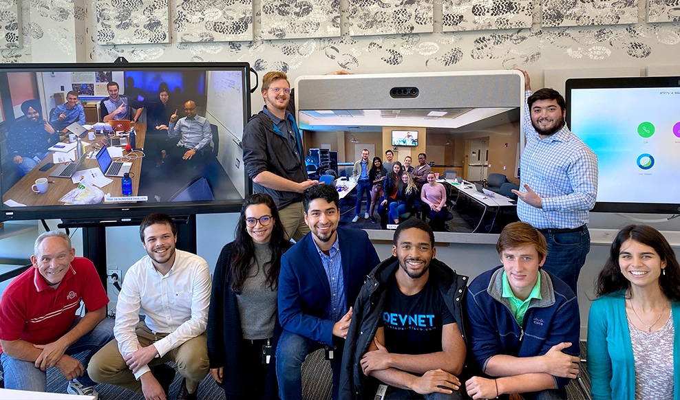 Group of nine people smiling with two videoconferences behind them.
