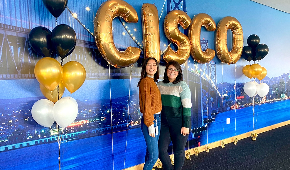 Two people pose together in front of balloons that spell out, 'Cisco.'