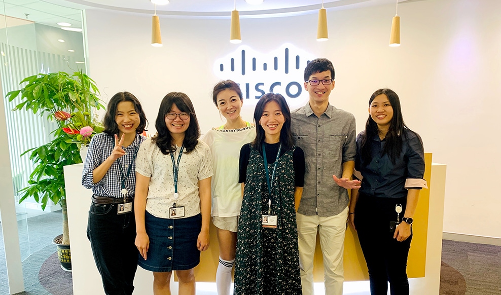 Group of six smiling people stand together with the Cisco logo in the background.