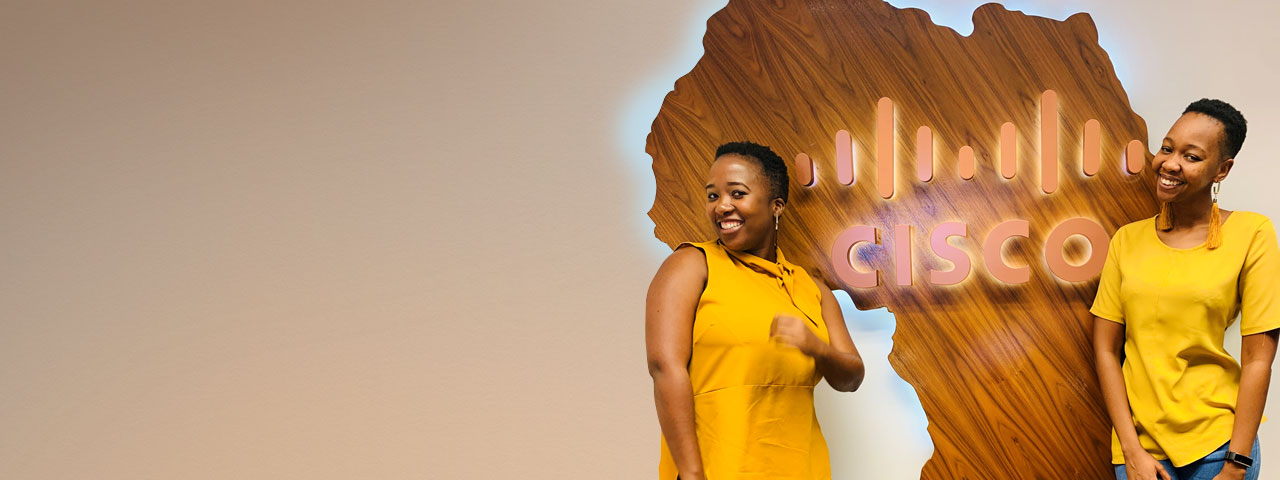 Two females wearing yellow in front of a brown wooden sign shaped like Africa with the Cisco Logo