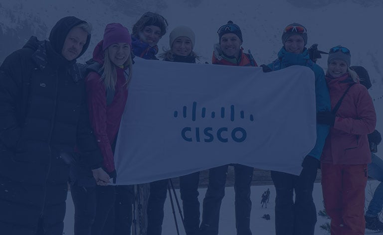 Seven employees dressed in winter gear holding a white Cisco sign with snow mountains in the background.