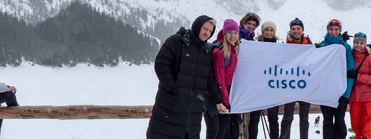 Seven employees dressed in winter gear holding a white Cisco sign with snow mountains in the background.