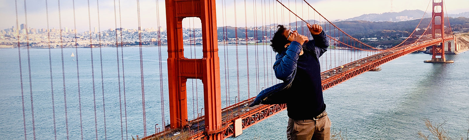 Male holding two thumbs up and looking to the sky with the Golden Gate Bridge and ocean in the background