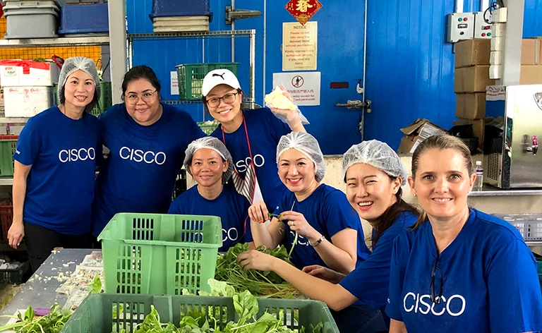Seven women wearing the same blue Cisco shirt volunteering at a food bank while sorting lettuce