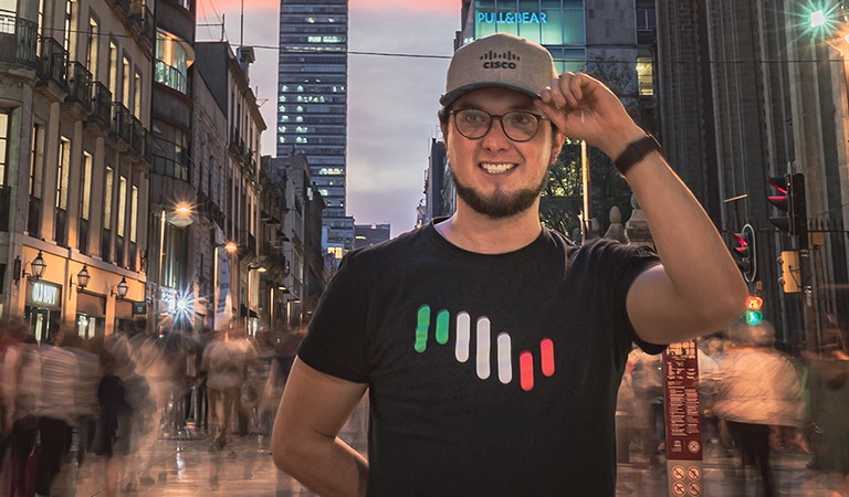 Male wearing a Cisco Hat and shirt holding left hand to his hat on a street in Mexico City