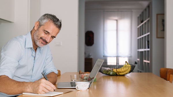 Mann sitzt in der Küche, macht Notizen, unterstützt von Technologie