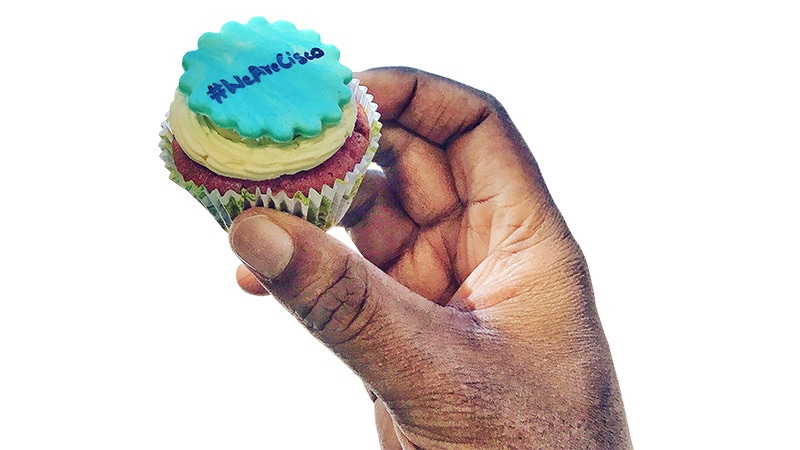 Hand holding a blue, yellow, and red cupcake that reads “#WeAreCisco.”