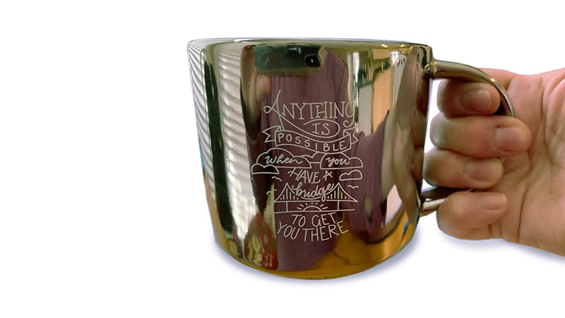 Hand holding a gold mug that reads, “Anything is possible when you have a bridge to get you there.”