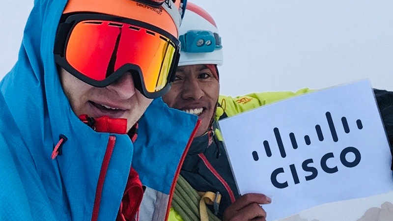 Two males on a snowy mountain in Bolivia, wearing goggles, rock climbing hats holding white paper with the Cisco Logo on it