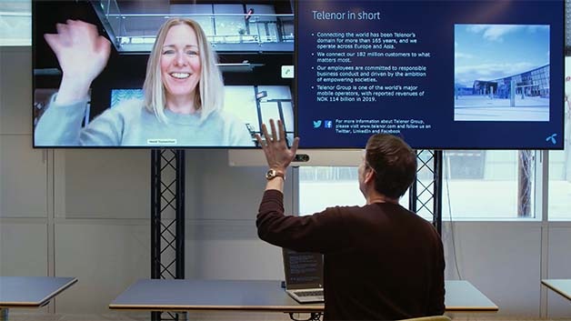 A woman on a large screen waves to a male colleague in an office via Cisco’s Webex solution