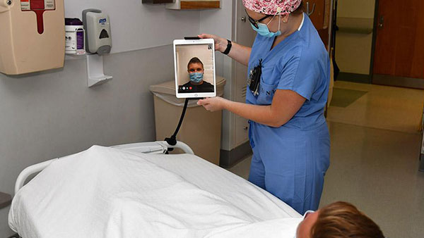 A nurse facilitating a conversation between a doctor and a patient lying in hospital bed via iPad 