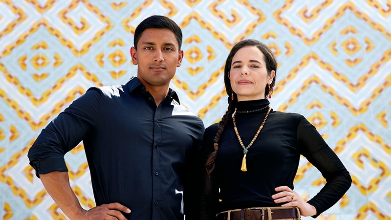 Two people standing in front of a wall mosaic looking at the camera with intention and purpose.