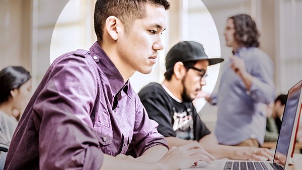 The side profile of a young man sitting in a class examining his laptop screen with his classmates and teacher in the background