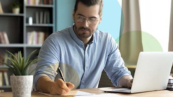 A bearded man wearing glasses in a blue shirt writing on a piece of paper in front of his laptop at home with a potted plant next to him on the desk