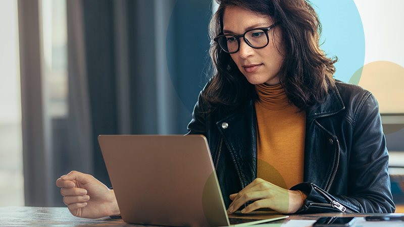 A woman with dark hair wearing glasses in a leather jacket and orange turtleneck sitting down in front of her laptop