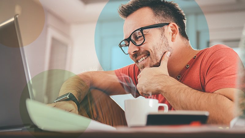 A man in an orange shirt and glasses smiling at his laptop working from home with a coffee cup next to him