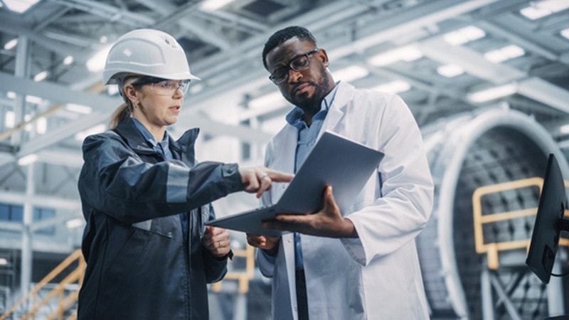 A worker wearing a hard hat pointing to a laptop held by a colleague in a white coat