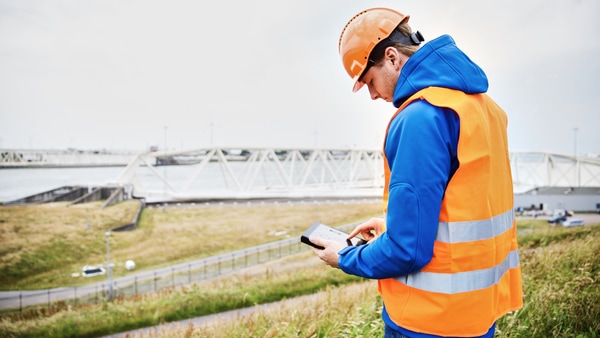 A worker wearing a hard hat and safety vest and holding a tablet in an outdoor industrial setting