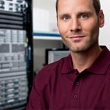 Portrait of male standing with a rack of routers and switches behind him