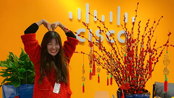 Female curling arms and placing hands on top of head to make a heart-shape against orange wall with Cisco logo affixed