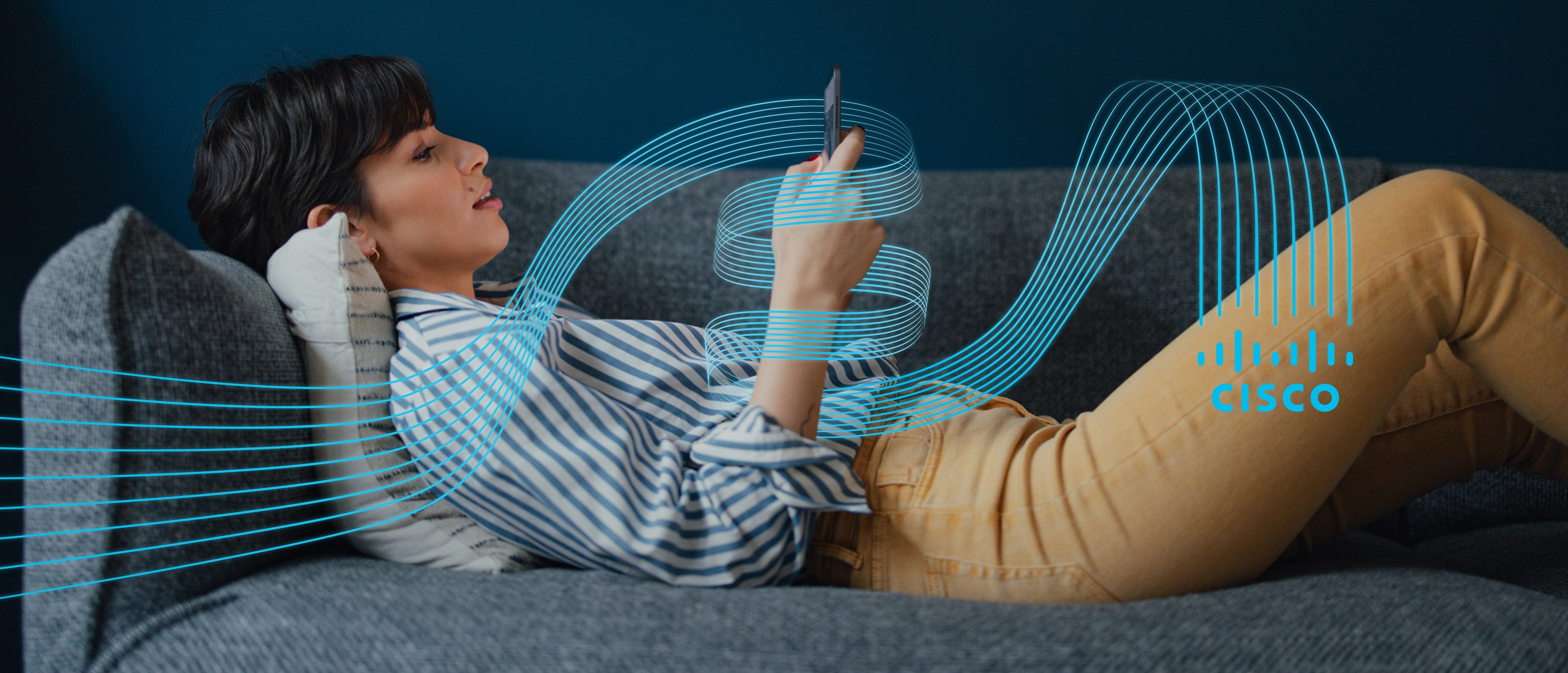 Woman laying on a gray couch in front of a dark blue wall holding cell phone wrapped in Cisco logo