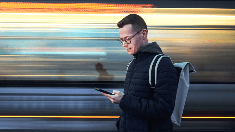 Image d’une personne avec un sac à dos regardant son téléphone portable tandis qu’un train semble passer à toute vitesse.