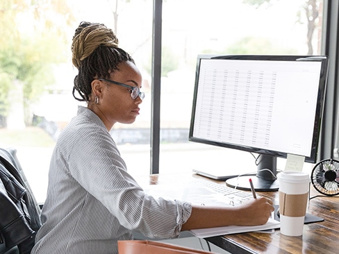 一名 IT 专业人员坐在办公桌前在活页笔记本上记笔记，其电脑屏幕上显示控制面板。